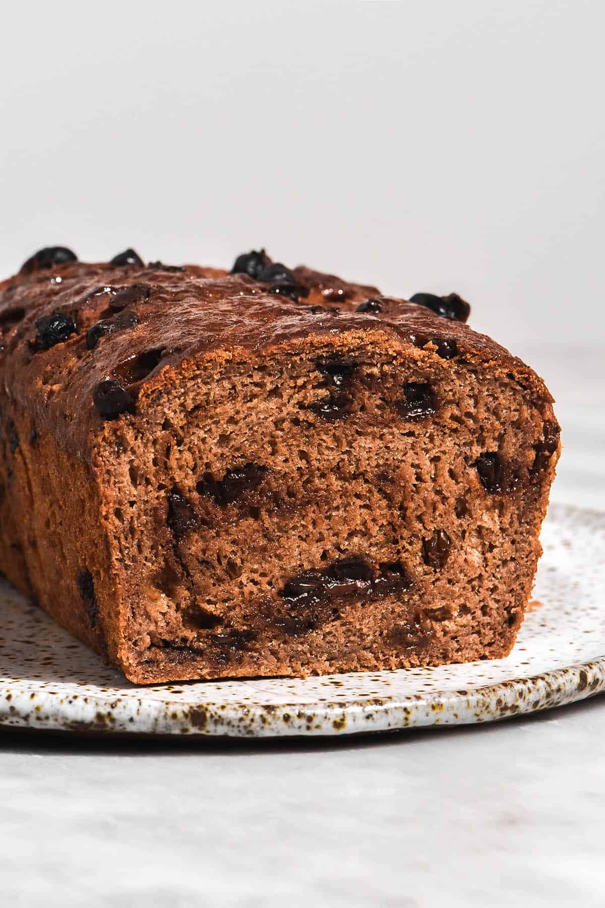 A brightly lit side on view of a gluten free hot cross bun loaf swirled with cinnamon sugar and studded with dark chocolate chips and raisins. The loaf has been sliced, revealing the crumb and melty chocolate inside. It sits on a white speckled ceramic plate against a white backdrop.