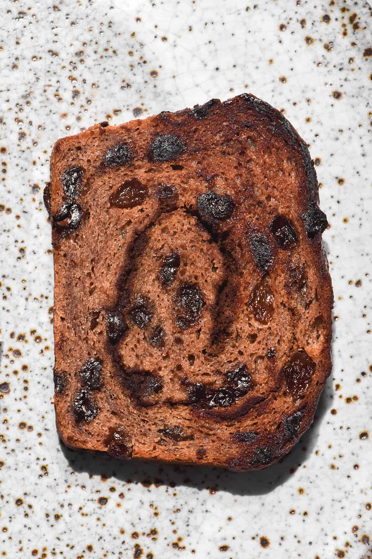 An aerial image of a slice of toasted gluten free hot cross bun loaf on a white speckled ceramic plate. The loaf is dotted with melted chocolate and raisins, and has a cinnamon swirl through the centre.