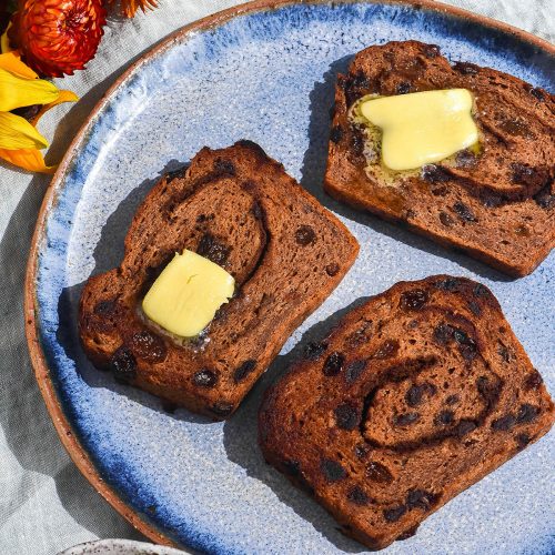 A brightly lit aerial view of a plate of gluten free hot cross bun loaf, sliced and toasted then topped with melting butter. The slices sit on a bright blue ceramic plate atop a pale green tablecloth. They are surrounded by brightly coloured flowers, a water glass and a ceramic dish filled with pastel Easter eggs.