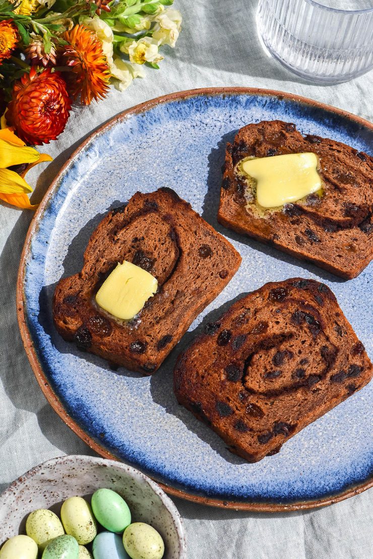 A brightly lit aerial view of a plate of gluten free hot cross bun loaf, sliced and toasted then topped with melting butter. The slices sit on a bright blue ceramic plate atop a pale green tablecloth. They are surrounded by brightly coloured flowers, a water glass and a ceramic dish filled with pastel Easter eggs.