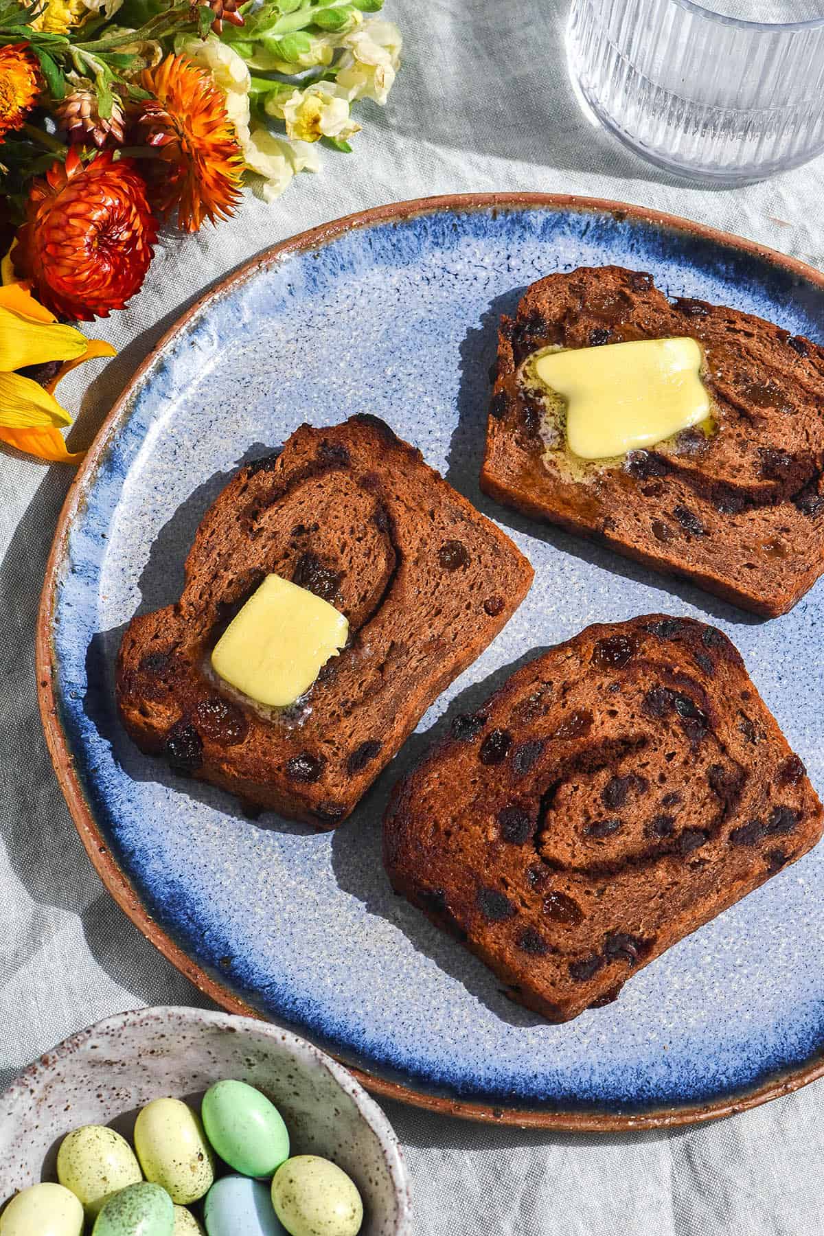 A brightly lit aerial view of a plate of gluten free hot cross bun loaf, sliced and toasted then topped with melting butter. The slices sit on a bright blue ceramic plate atop a pale green tablecloth. They are surrounded by brightly coloured flowers, a water glass and a ceramic dish filled with pastel Easter eggs. 