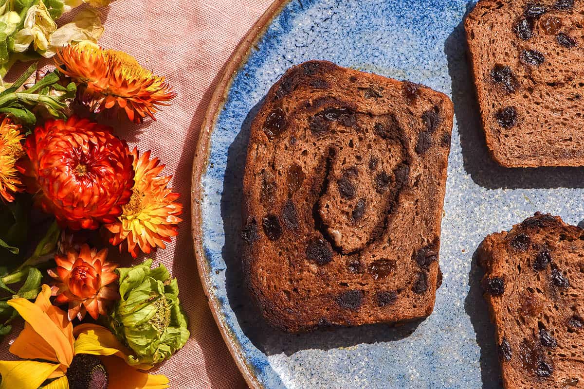 A bright blue ceramic plate topped with gluten free vegan cinnamon raisin loaf slices. The plate sits atop a pale pink linen tablecloth and brightly coloured flowers sit to the left of the image