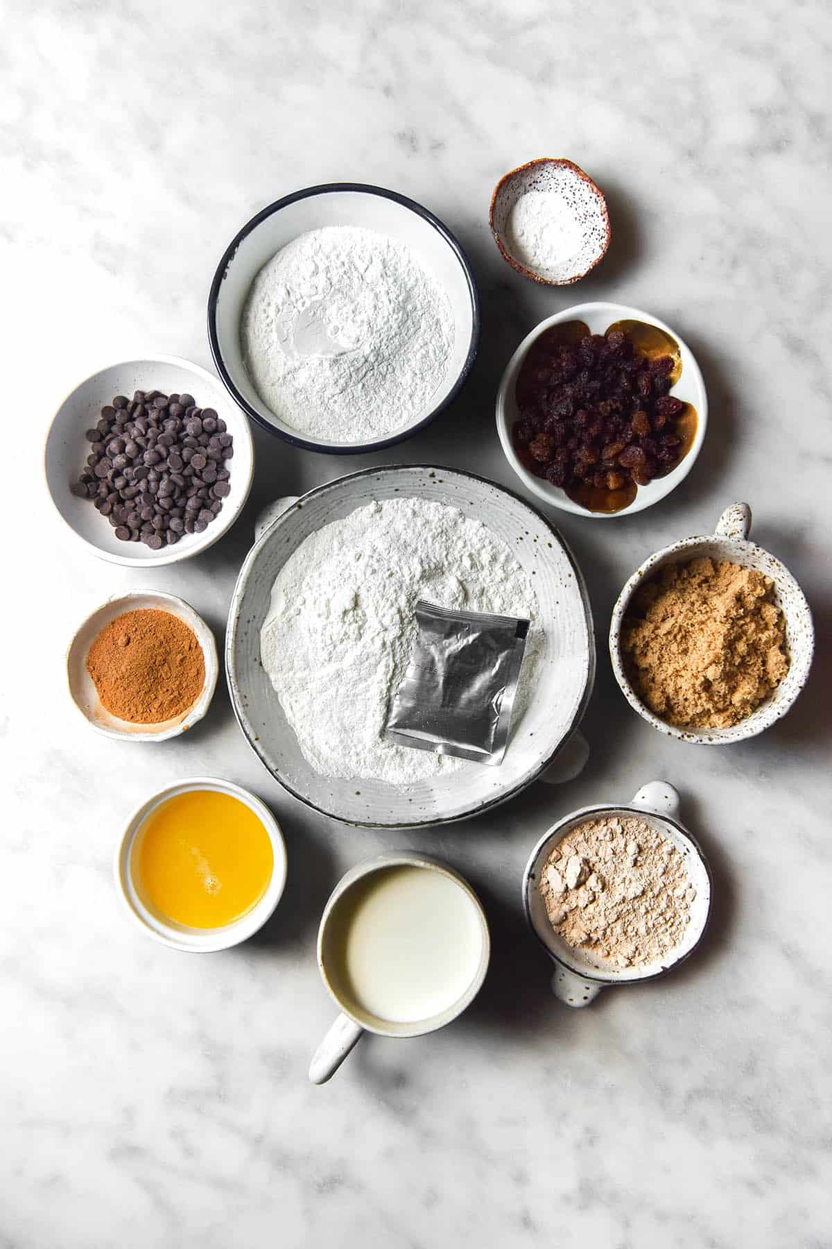 An aerial image of the ingredients used to make a gluten free hot cross bun loaf. The ingredients are arranged in small white ceramic bowls on a brightly lit white marble table