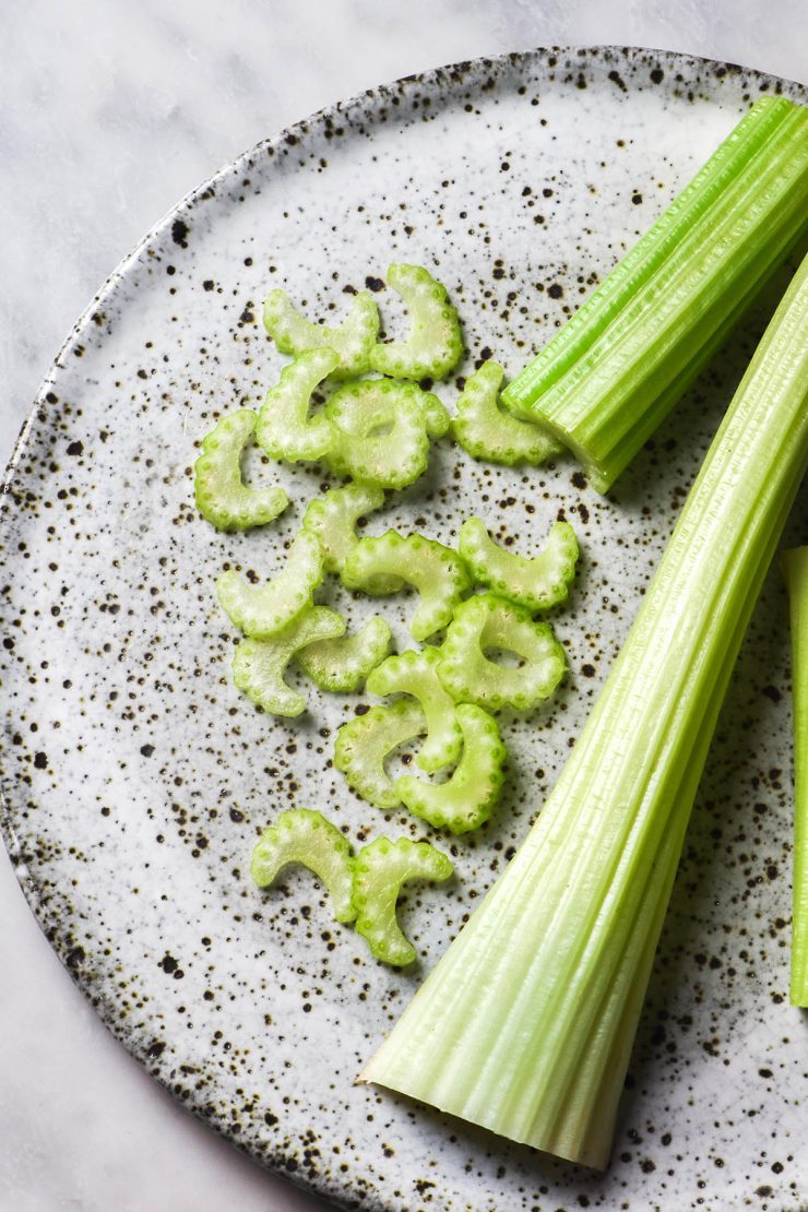 An aerial image of a white speckled ceramic plate topped with one stalk of celery and another that has been half sliced, with the half moon slices casually arranged on the plate. The plate sits atop a white marble table.