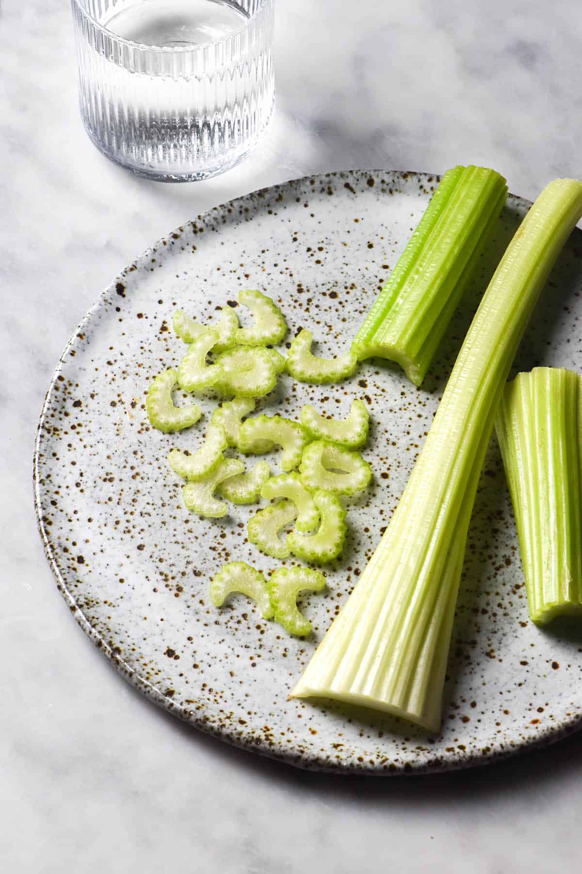 An aerial, side on image of two stalks of celery and some celery slices on a white speckled ceramic plate atop a white marble table. A textured water glass sits to the top left of the image.