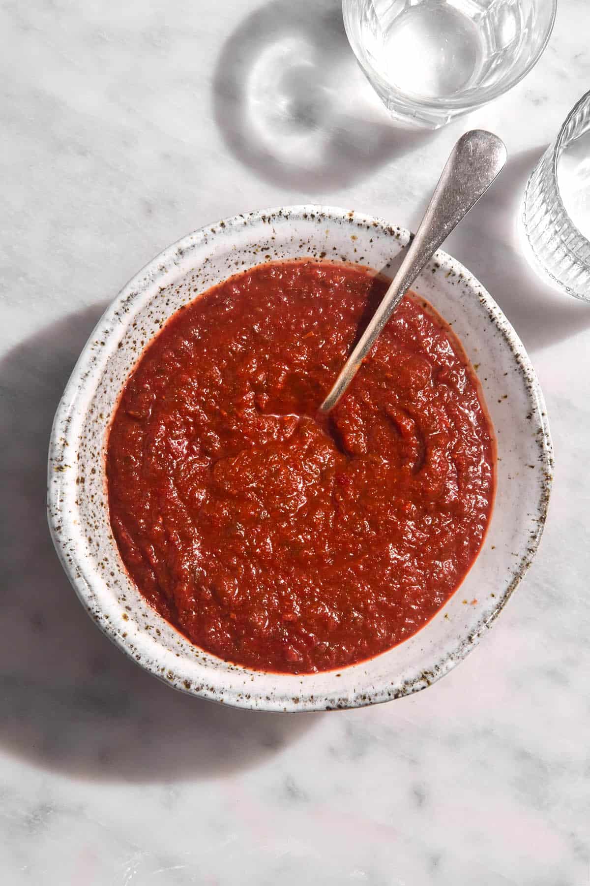 An aerial image of a white speckled ceramic bowl filled with low FODMAP Nomato sauce. The bowl sits on a white marble in bright sunlight and two glasses of water sit in the top right corner.