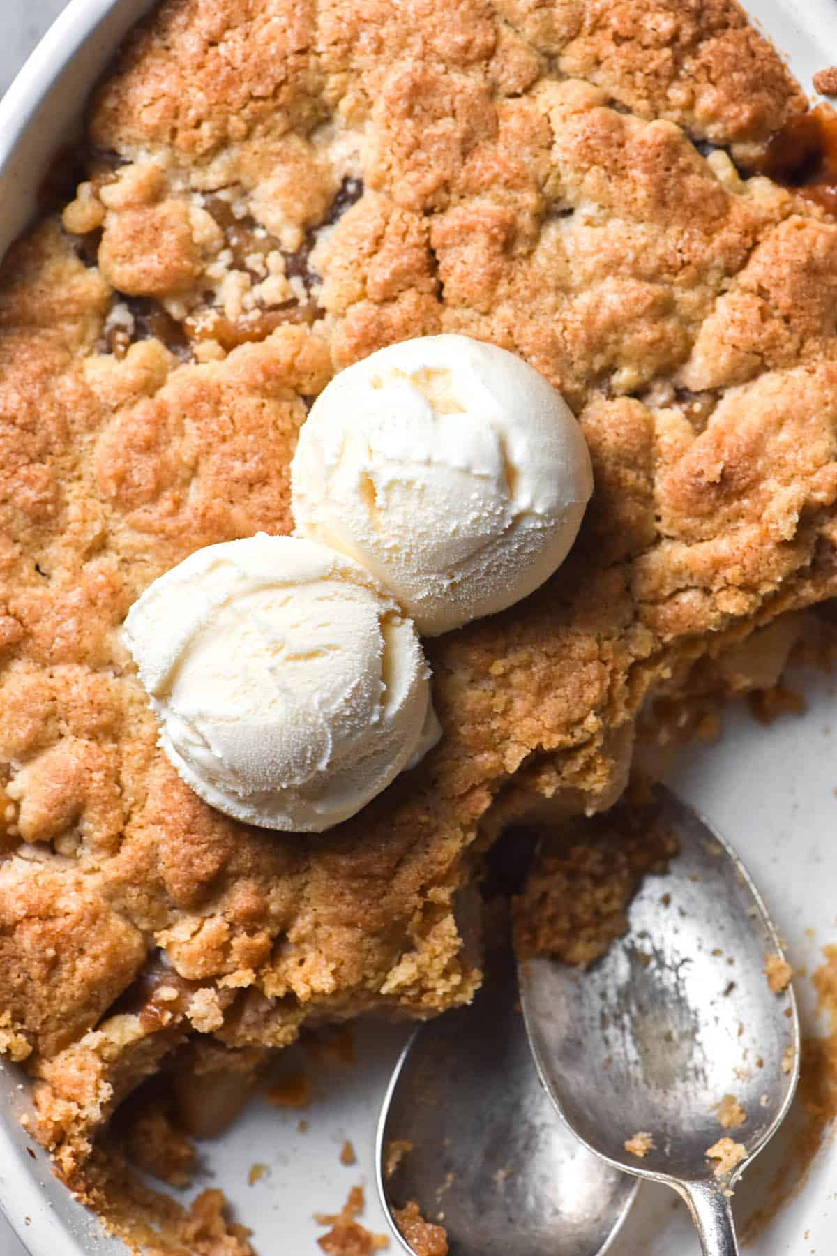 An aerial image of a low FODMAP apple pie in a white ceramic baking dish. Part of the crumble has been eaten, and two spoons sit in the empty space. Two scoops of vanilla ice cream sit in the middle of the crumble.