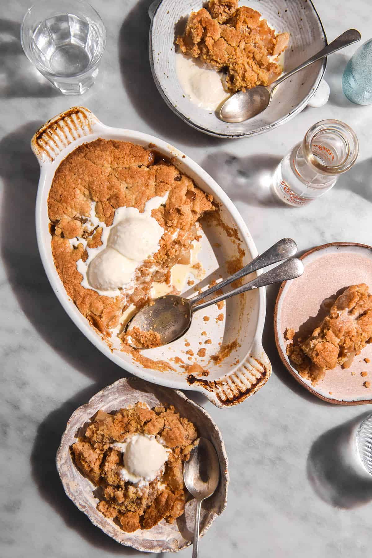 An aerial image of a low FODMAP apple crumble in a white ceramic baking dish on a white marble table. The crumble is topped with two scoops of vanilla ice cream, and has had a few servings removed and two spoons sit in the empty space. The baking dish is surrounded by bowls of crumble topped with vanilla ice cream. 
