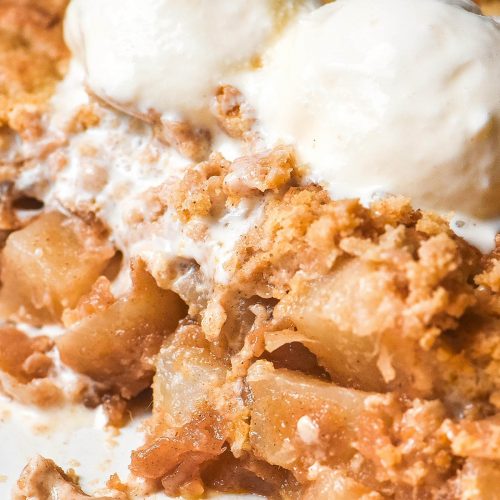 A close up image of a low FODMAP apple crumble in a white ceramic baking dish. Some crumble has been removed, revealing the cubes of 'apple' underneath. The crumble is topped with two scoops of vanilla ice cream.