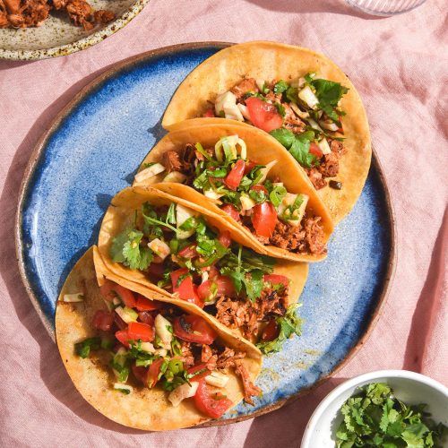 An aerial image of low FODMAP jackfruit tacos topped with pico de gallo on a bright blue ceramic plate. The plate sits atop a pale pink linen tablecloth/