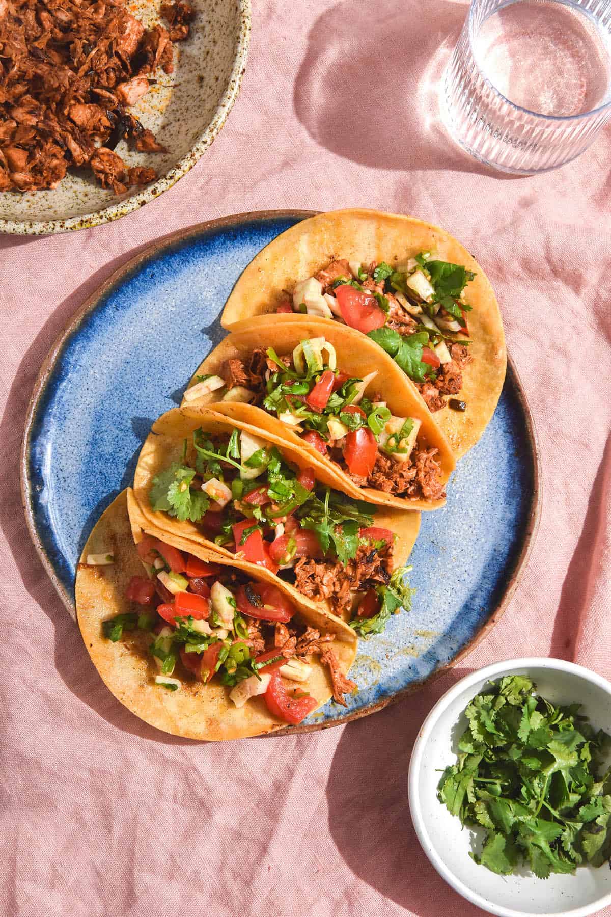 An aerial brightly lit view of a blue ceramic plate topped with low FODMAP jackfruit tacos sits on a pale pink linen tablecloth. A glass of water sits in the top right of the image, while a small white bowl of coriander sits in the bottom right of the image. 