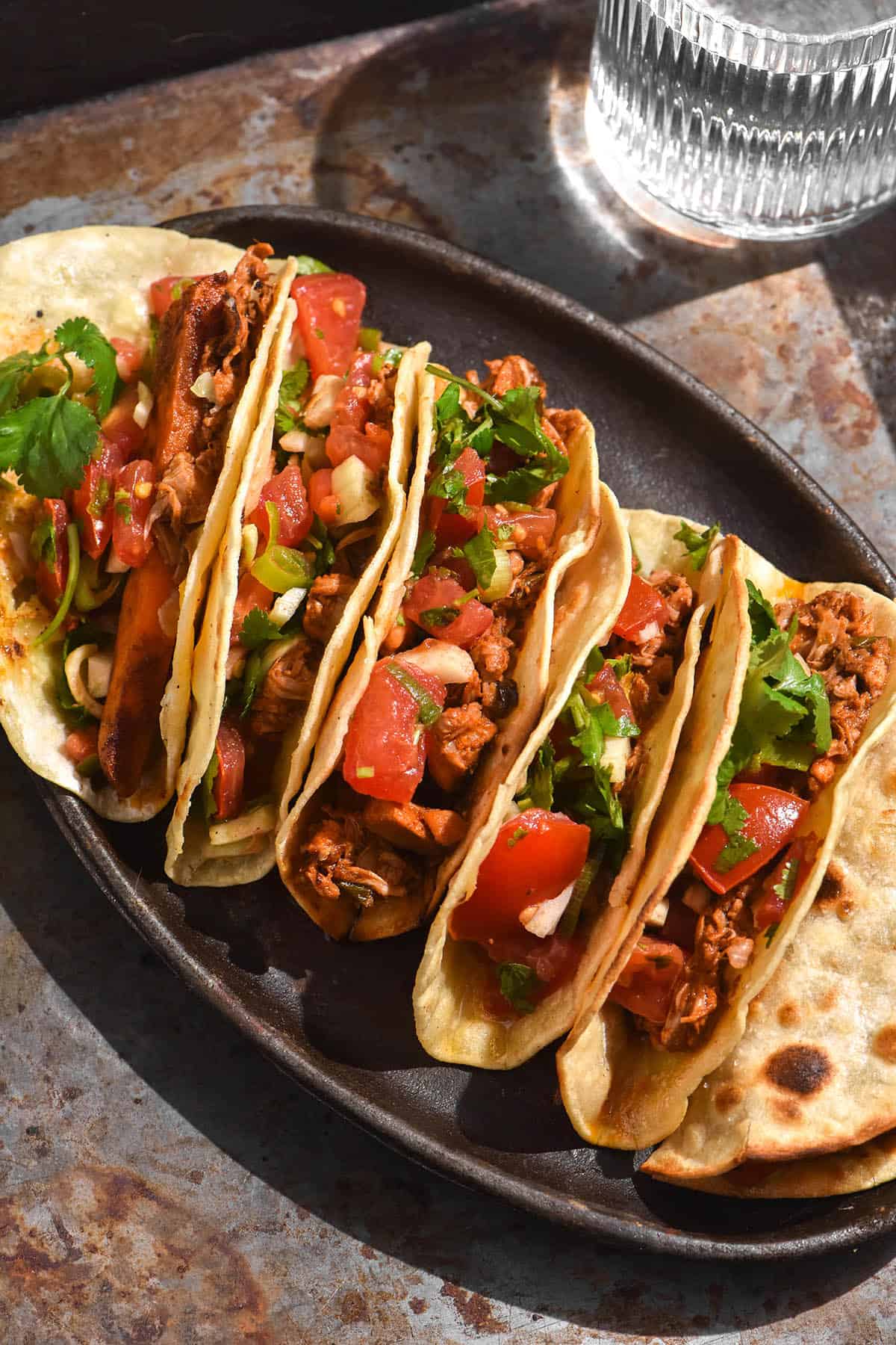A side on image of low FODMAP tacos filled with pico de gallo on a steel sizzler platter against a steel backdrop. A glass of water sits in the background.