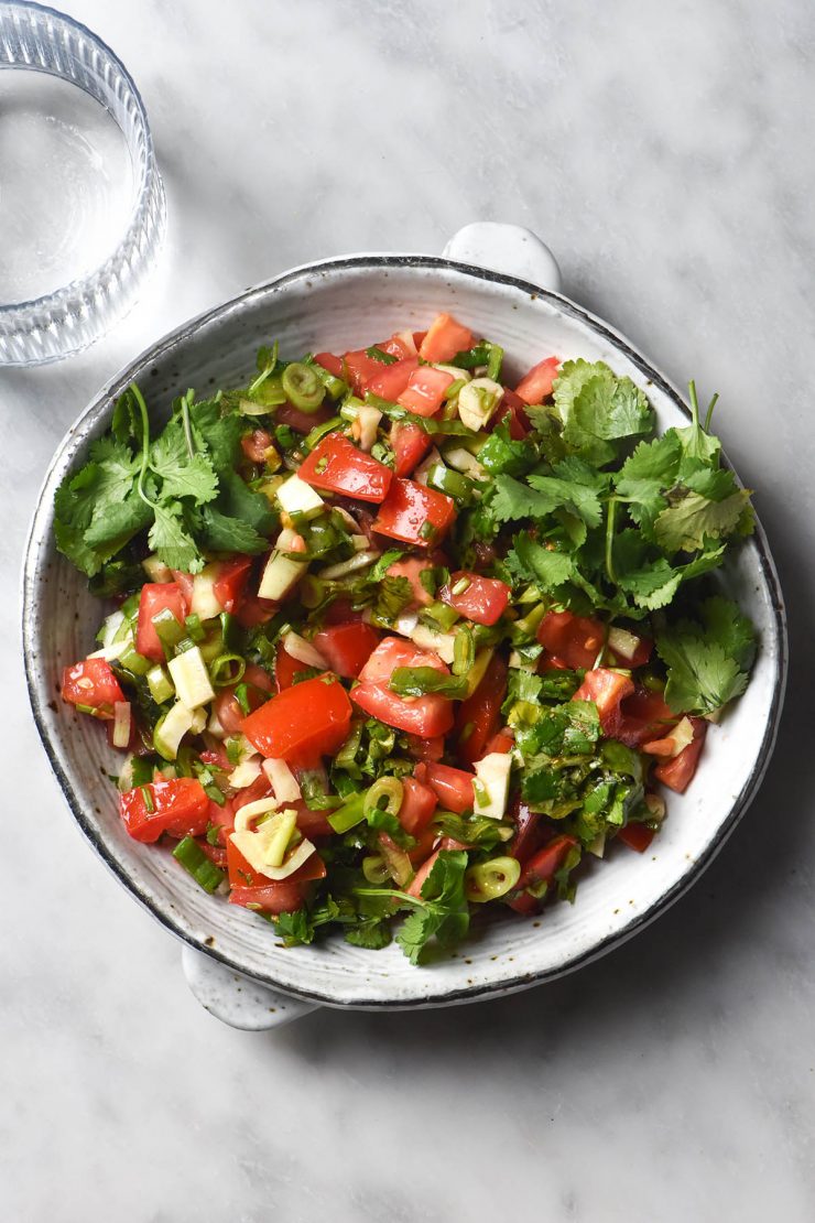 An aerial image of a white ceramic plate filled with low FODMAP pico de gallo sits on a white marble table. A glass of water sits to the top left of the image.