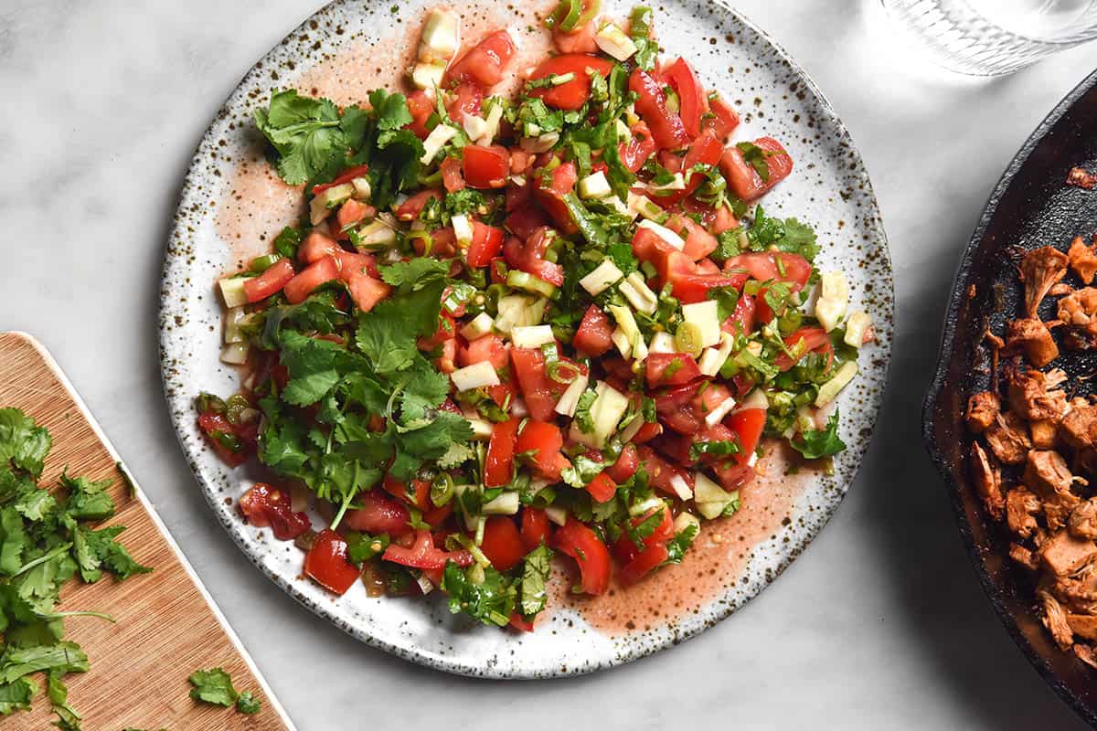 An aerial image of a white speckled ceramic plate of low FODMAP pico de gallo on a white marble table. Jackfruit mince and a chopping board topped with coriander surround the plate. 