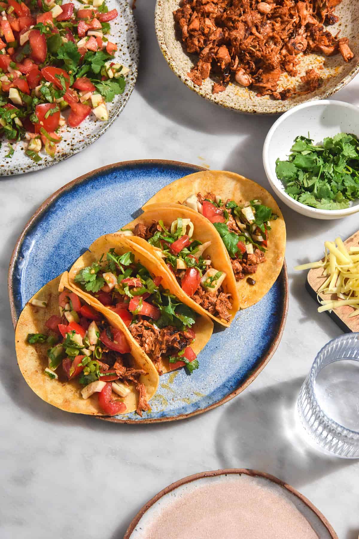 An aerial brightly lit view of a white marble table topped with taco ingredients. A plate of tacos sits in the centre and a low FODMAP pico de gallo sits to the top left of the image, and jackfruit taco mince sits the top right. Water glasses, extra condiments and plates sit casually strewn around the edges of the image