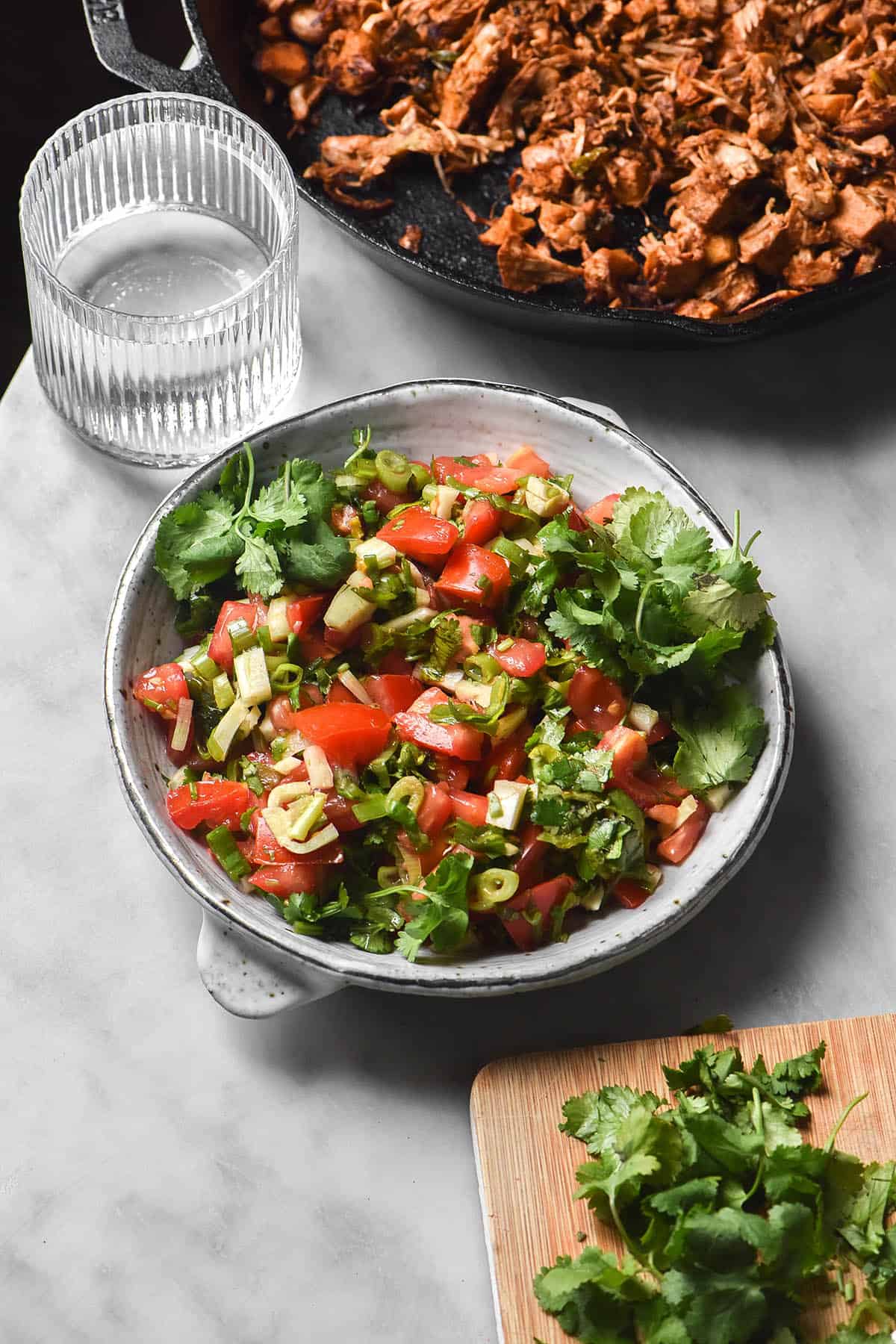 A brightly lit image of a white ceramic bowl filled with low FODMAP pico de gallo on a white marble table. A skillet filled with jackfruit taco mince and a water glass sit in the background, while a chopping board topped with coriander sits in the foreground