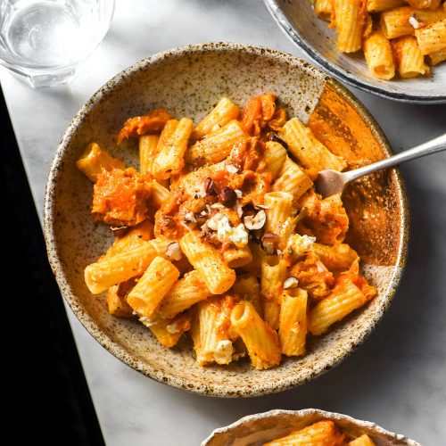 An aerial image of white ceramic plates filled with low FODMAP pumpkin pasta on a white marble table. The pasta is topped with goat's cheese and chopped hazelnuts.