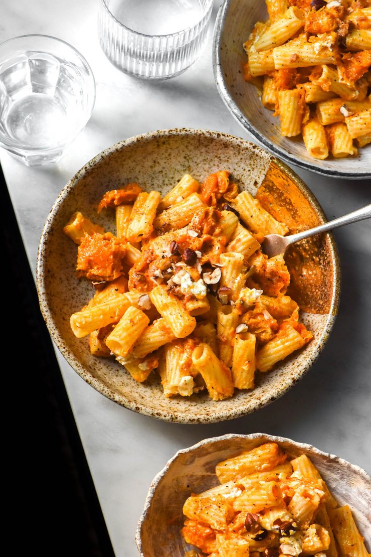 An aerial image of white ceramic plates filled with low FODMAP pumpkin pasta on a white marble table. The pasta is topped with goat's cheese and chopped hazelnuts.