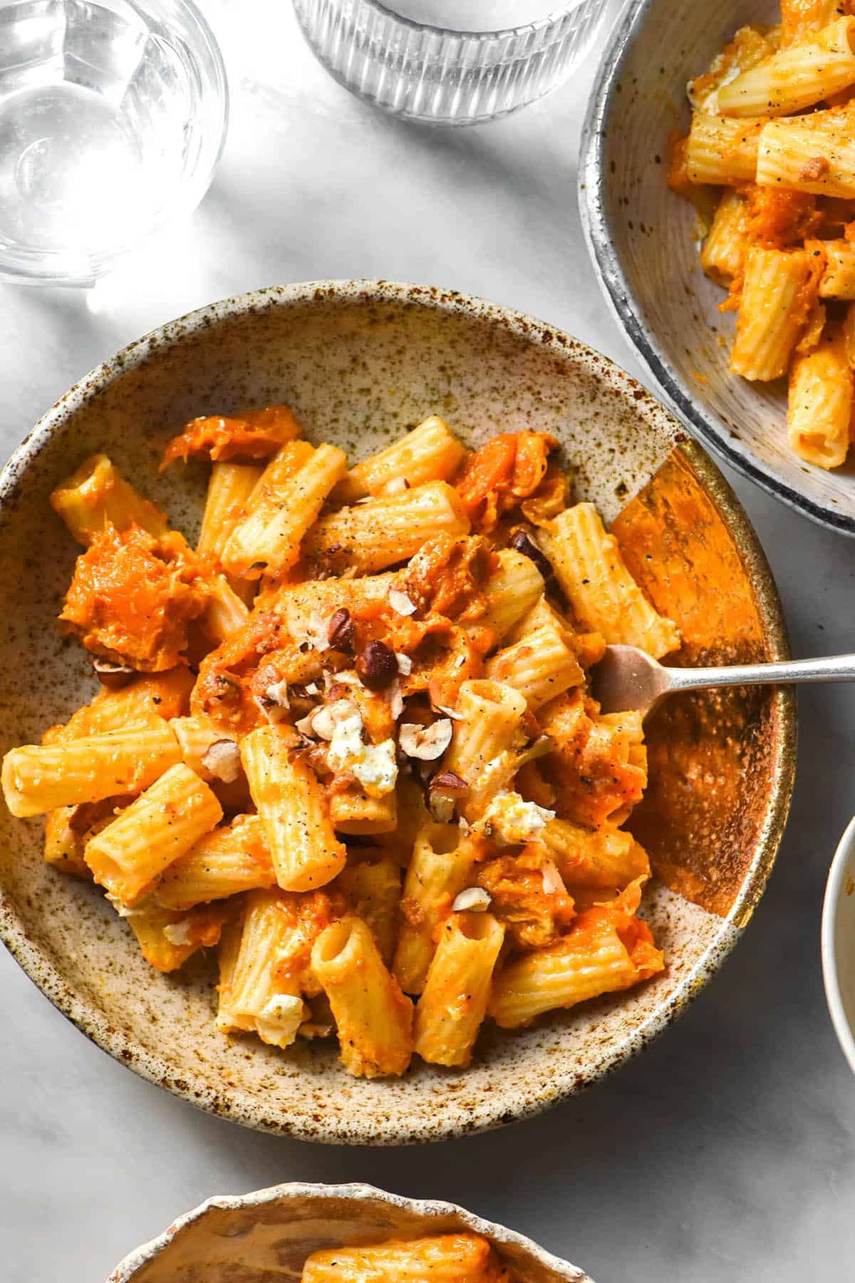 An aerial image of white ceramic plates filled with low FODMAP pumpkin pasta on a white marble table. The pasta is topped with goat's cheese and chopped hazelnuts. 