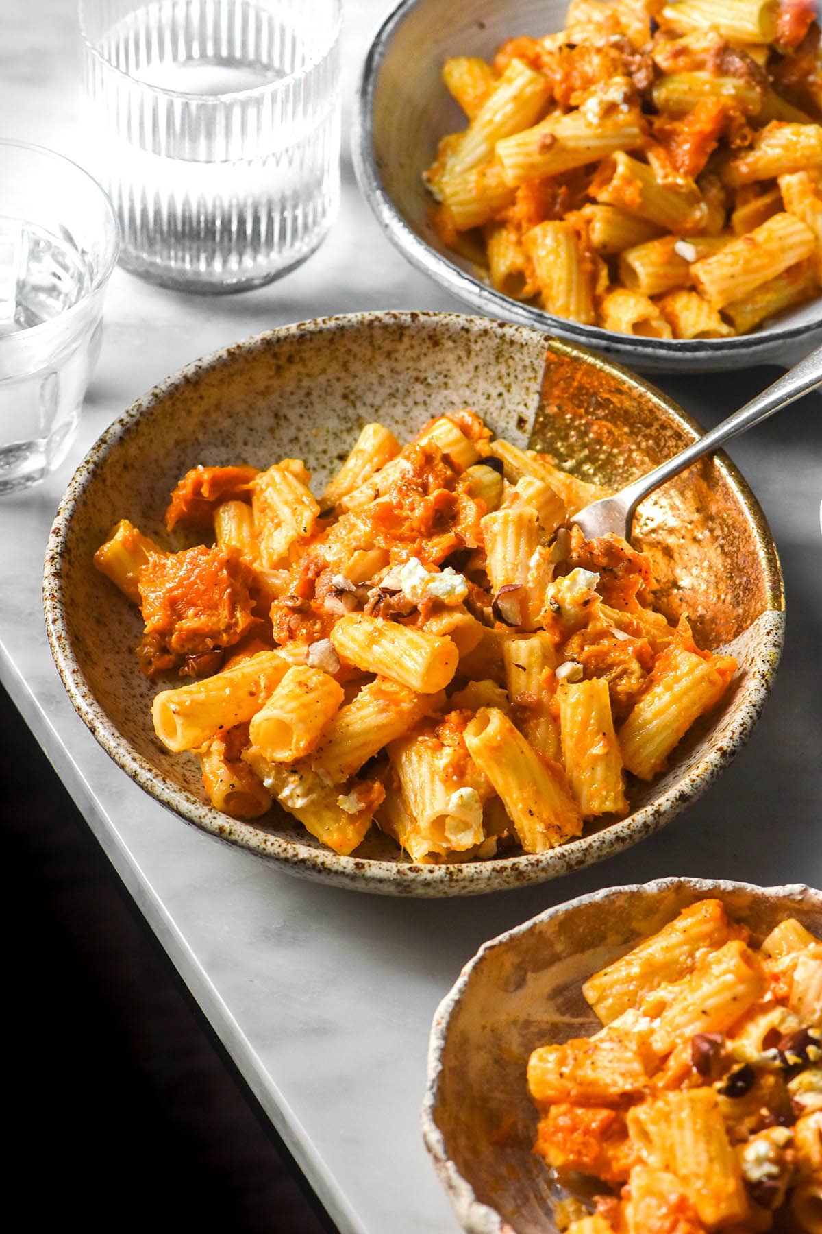 An aerial image of white ceramic plates filled with low FODMAP pumpkin pasta on a white marble table. The pasta is topped with goat's cheese and chopped hazelnuts. 