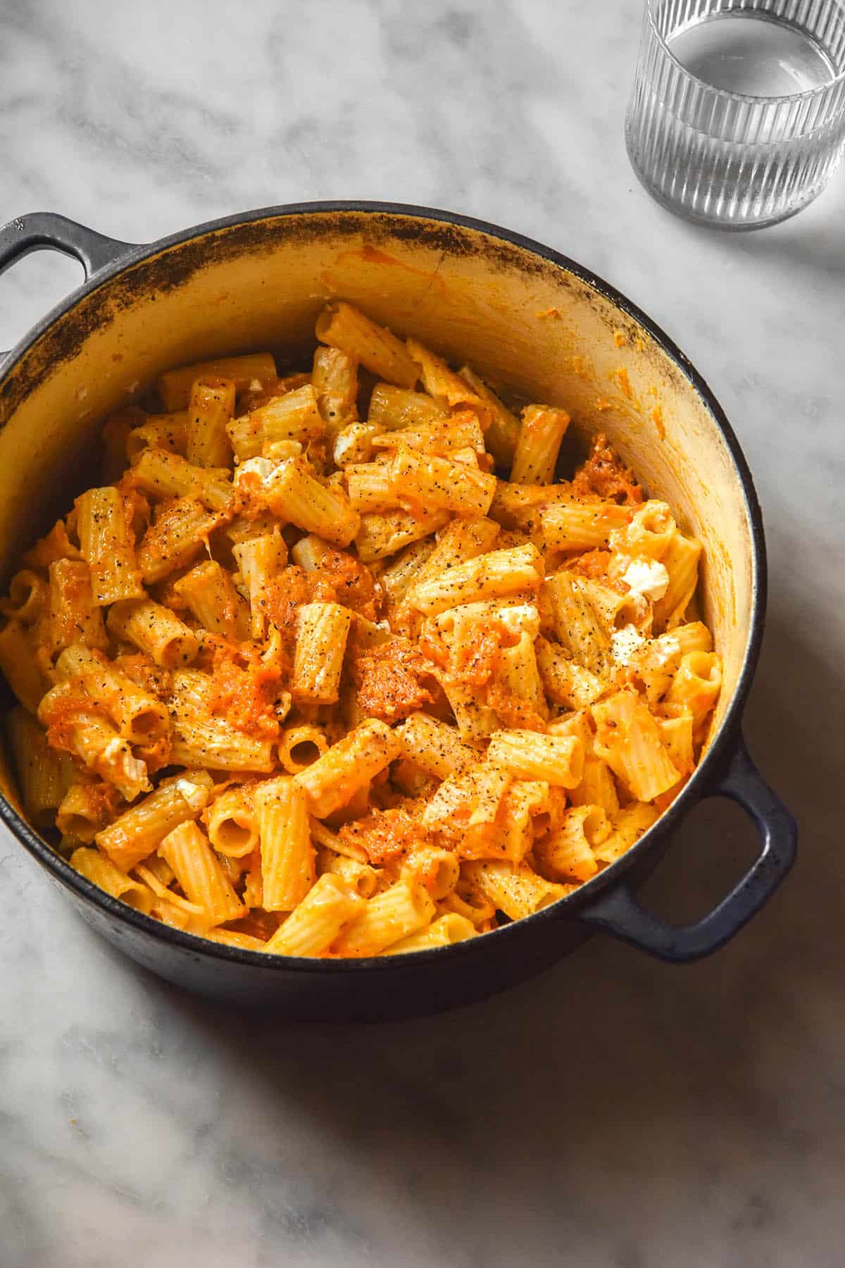 An aerial image of a Dutch oven filled with low FODMAP pumpkin and goat's cheese pasta. The Dutch oven sits on a white marble table and a water glass sits to the top right of the image. 