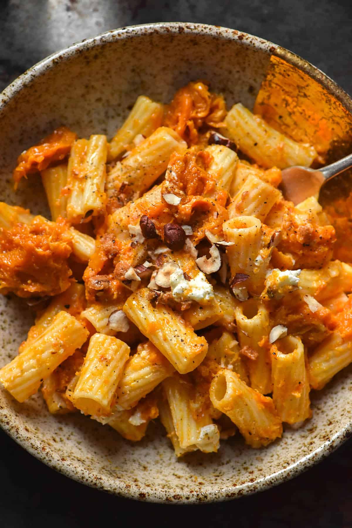 An aerial close up image of a bowl of pumpkin and goat's cheese rigatoni topped with chopped hazelnuts. The pasta sits in a beige speckled ceramic bowl on a dark steel backdrop.