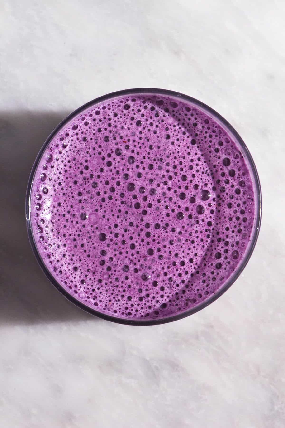 An aerial image of a frothy blueberry smoothie on a white marble table in harsh sunlight
