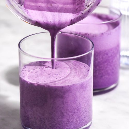 A brightly lit image of two glasses of bright purple blueberry smoothie. A blender pours more smoothie into the cup in the foreground.