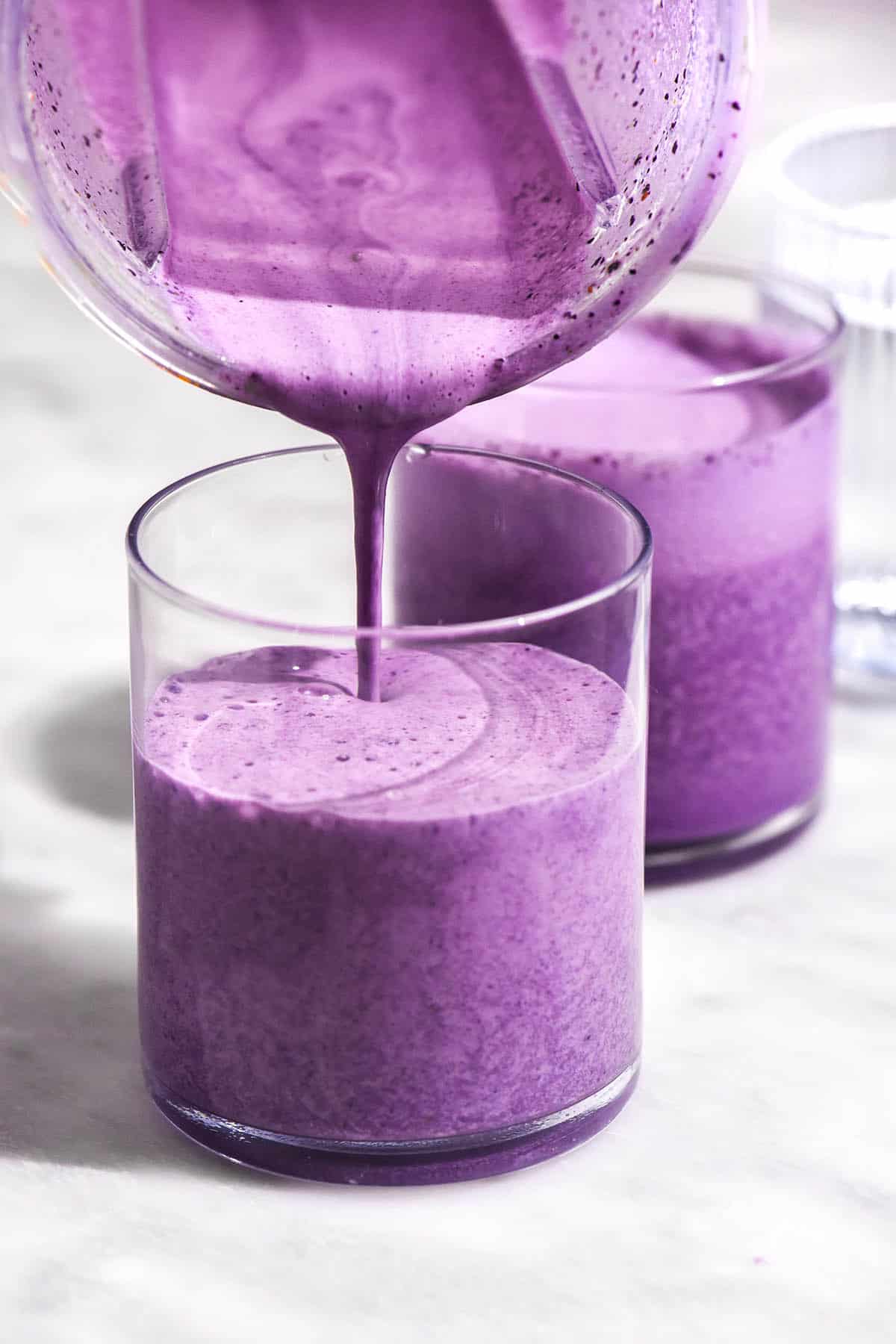 A brightly lit image of two glasses of bright purple blueberry smoothie. A blender pours more smoothie into the cup in the foreground.