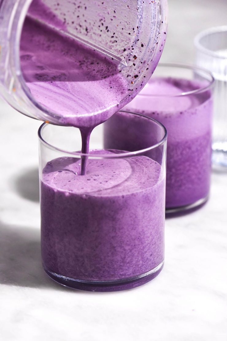 A brightly lit image of two glasses of bright purple blueberry smoothie. A blender pours more smoothie into the cup in the foreground.