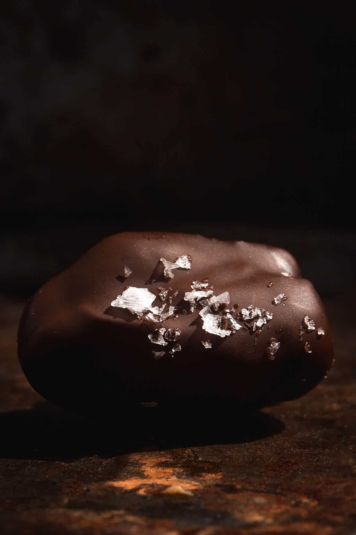 A dark and moody side on image of a Medjool date coated in chocolate and sprinkled with sea salt flakes