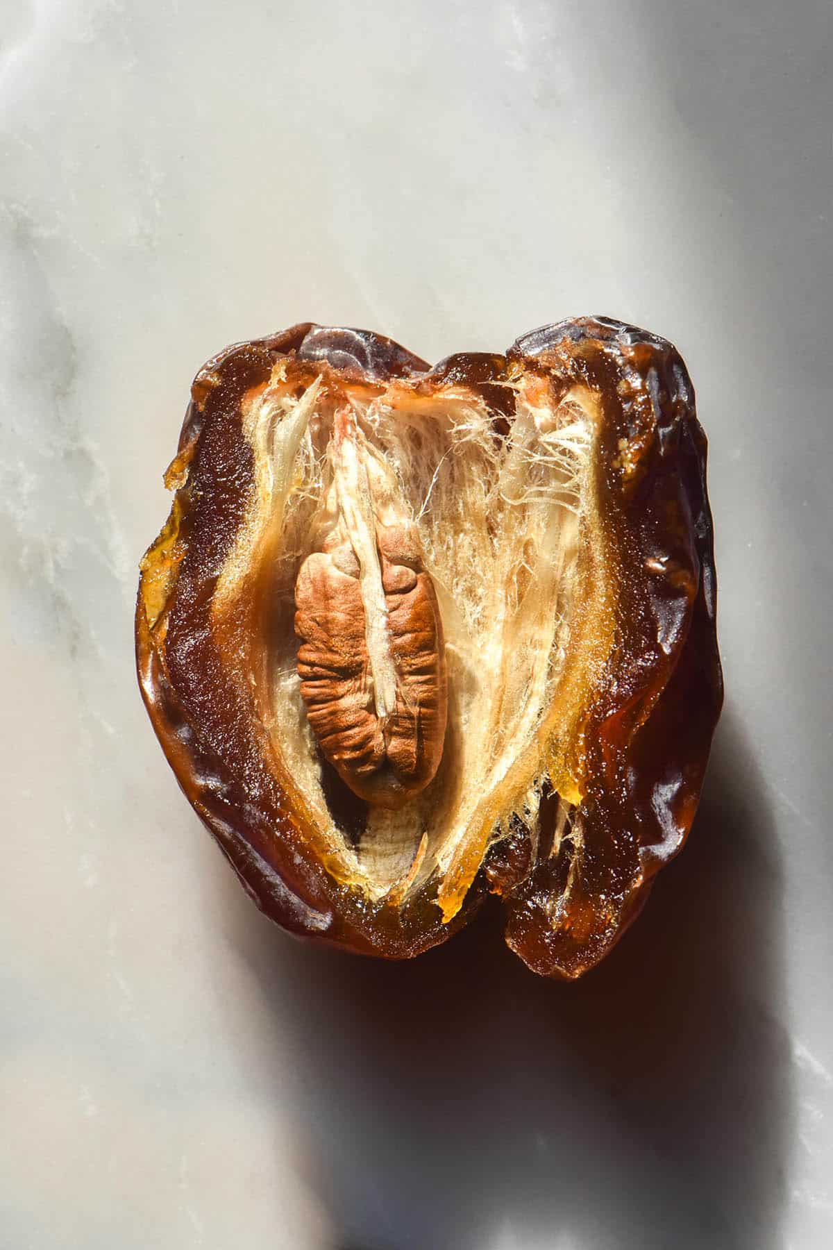 A macro aerial close up image of a Medjool date that has been sliced open to reveal the seed and the insides of the date. The date sits atop a white marble table in sunlight. 
