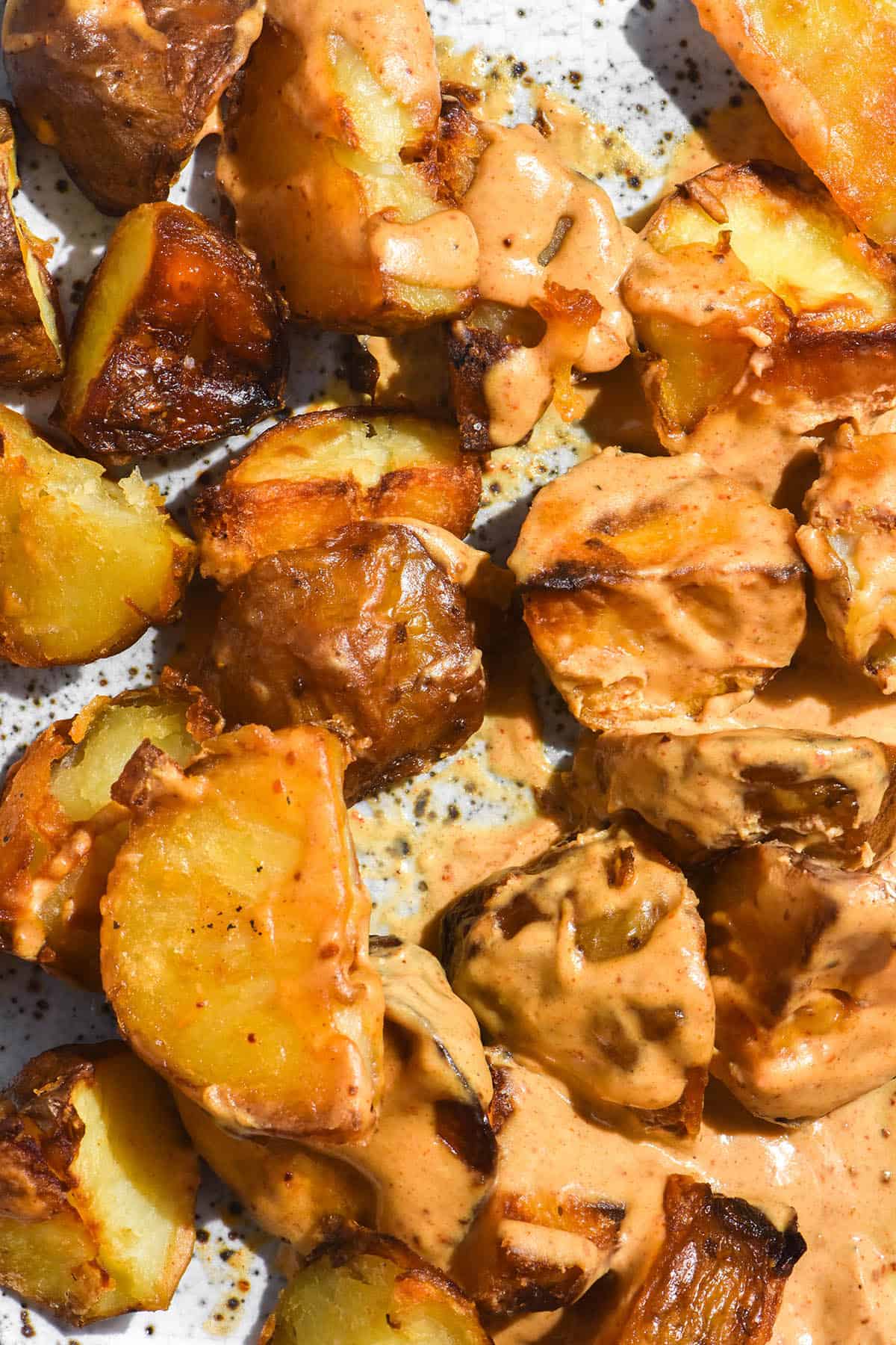 A brightly lit aerial image of crispy potatoes smothered in a low FODMAP smoky red tahini dressing. The potatoes sit on a white ceramic speckled plate. 