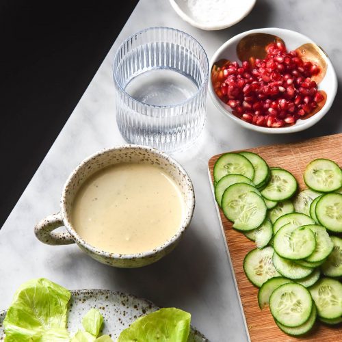 An aerial view of a low FODMAP tahini salad dressing in a white ceramic mug on a white marble table. The dressing is surrounded by ingredients that will be used in the salad.