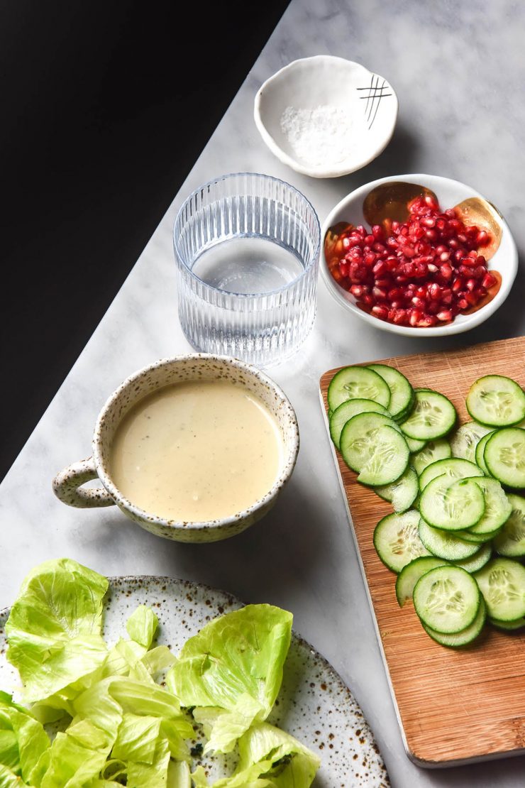 An aerial view of a low FODMAP tahini salad dressing in a white ceramic mug on a white marble table. The dressing is surrounded by ingredients that will be used in the salad.