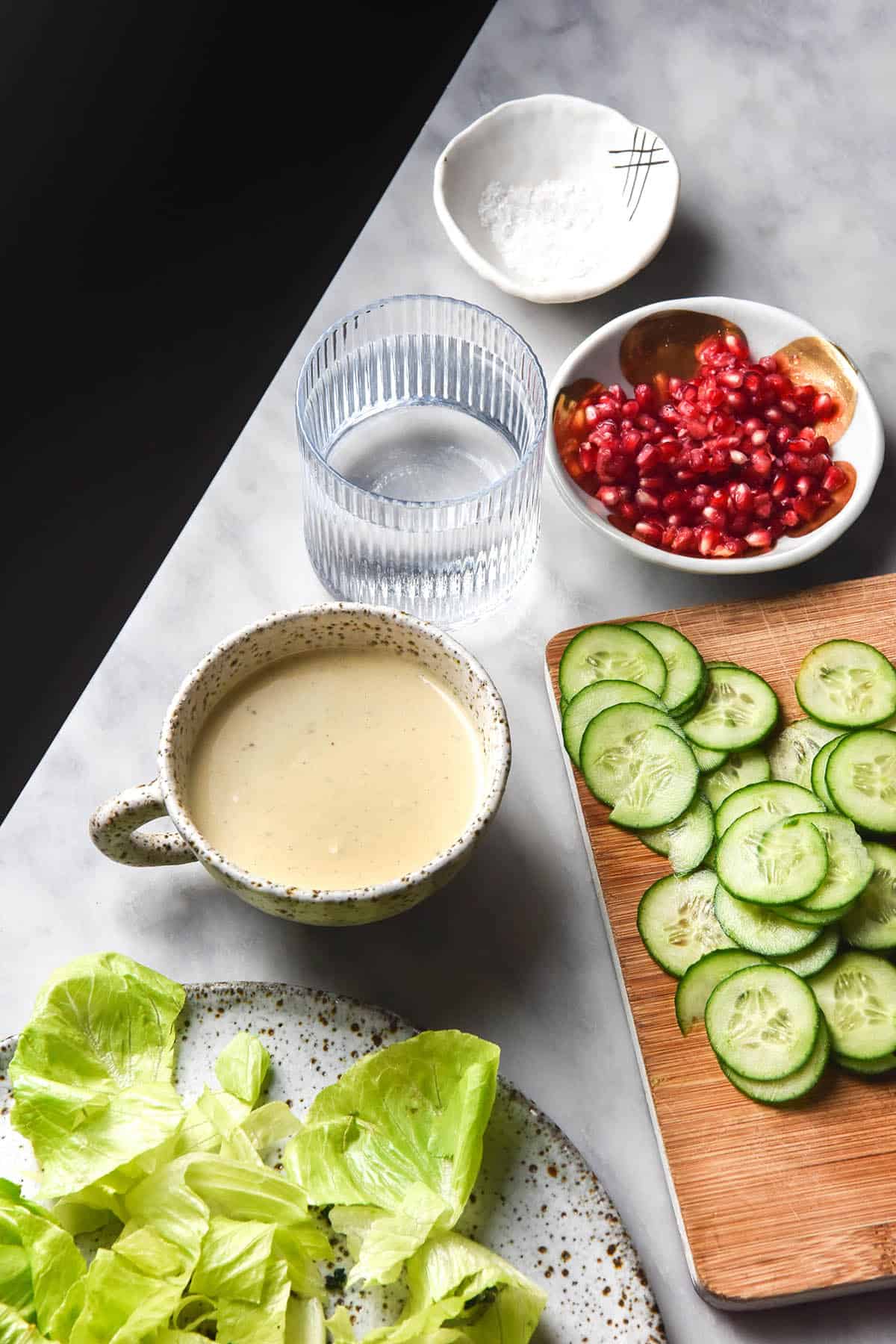 An aerial view of a low FODMAP tahini salad dressing in a white ceramic mug on a white marble table. The dressing is surrounded by ingredients that will be used in the salad. 