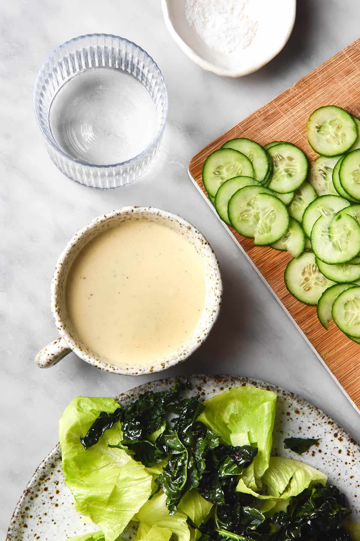 An aerial view of a low FODMAP tahini salad dressing in a white ceramic mug on a white marble table. The dressing is surrounded by ingredients that will be used in the salad.