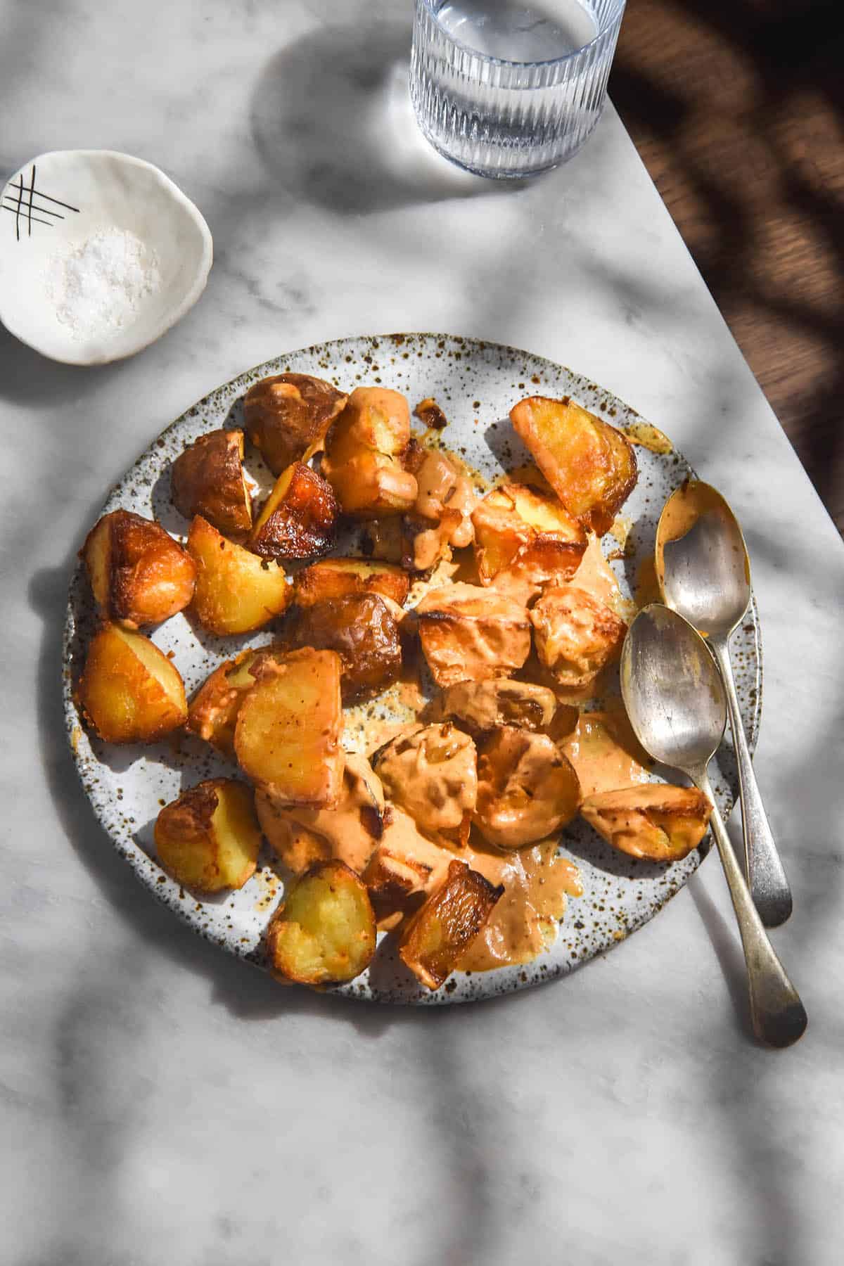 An aerial image of crispy potatoes smothered in a smoky orange tahini sauce. The potatoes sit on a white speckled ceramic plate against a white marble table in dappled light. 