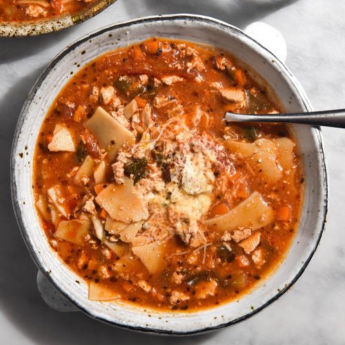 An aerial image of two white ceramic bowls filled with low FODMAP vegetarian lasagne soup. The lasagne soup is a deep red colour and topped with plenty of fresh vegetarian parmesan