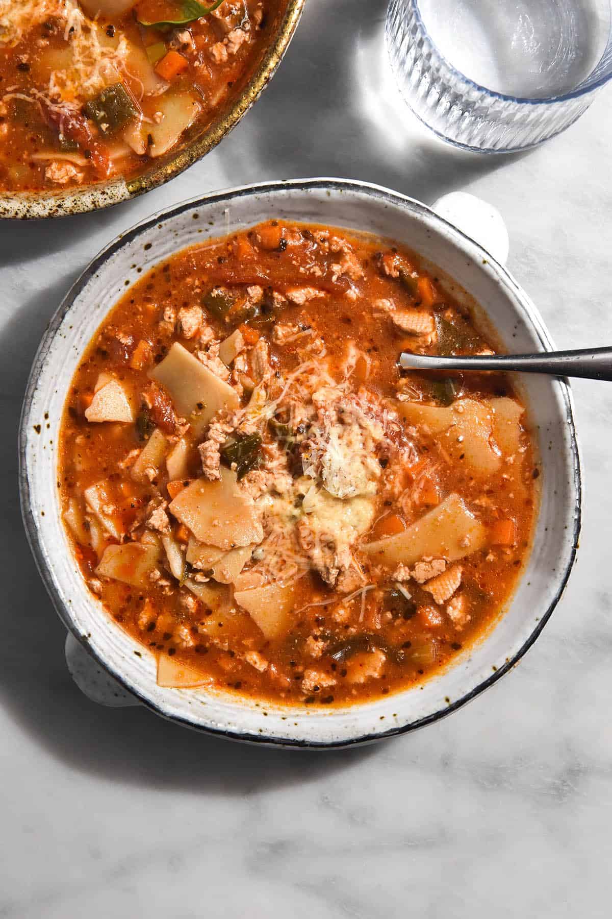 A brightly lit aerial image of a white speckled ceramic bowl of low FODMAP vegetarian lasagne soup on a white marble table. A spoon pokes into the bowl from the right hand side. A second bowl sits in the top left corner and a glass of water sits in the right hand corner. 