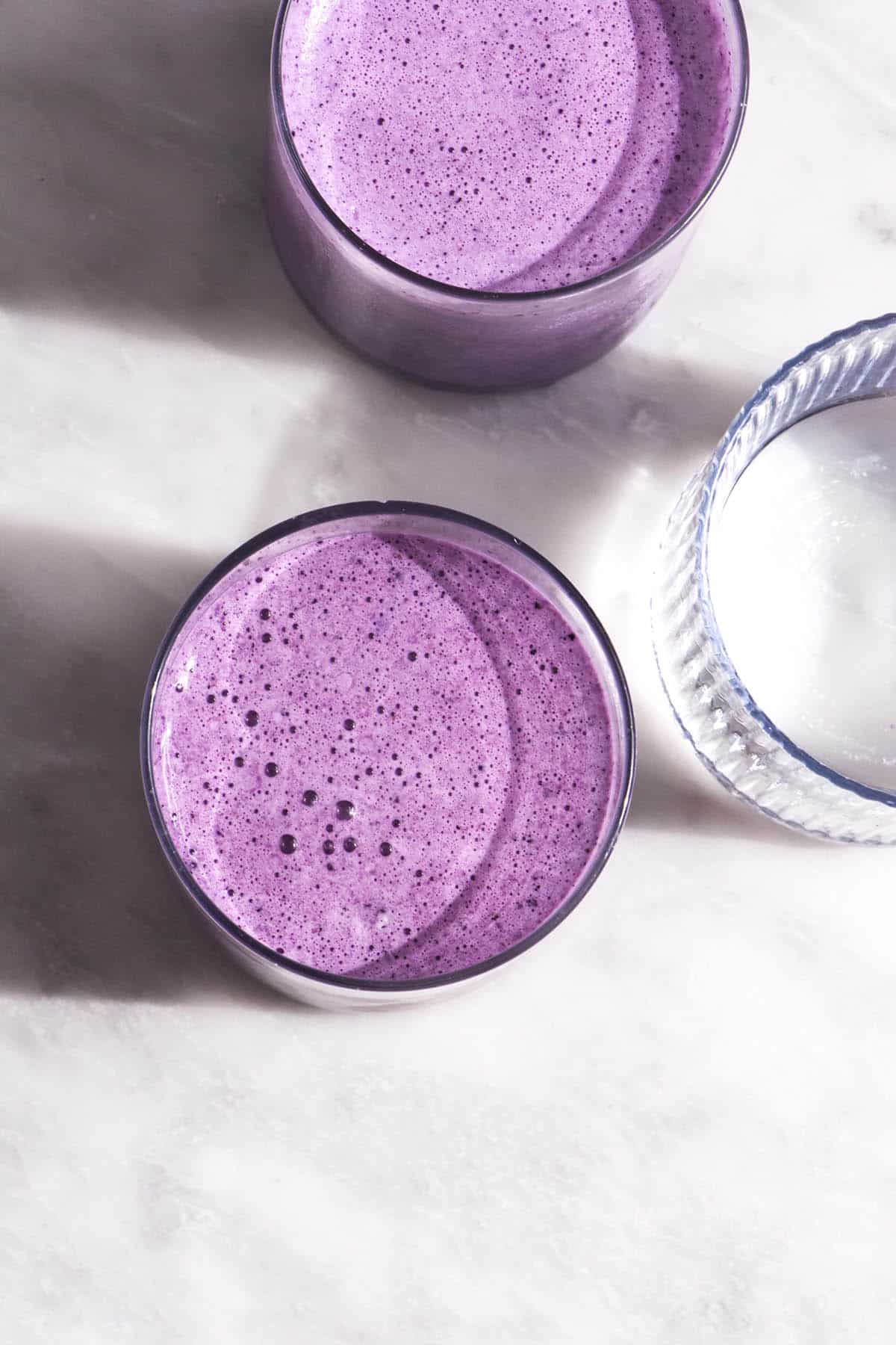 A brightly lit aerial image of two glasses of blueberry smoothie on a white marble table. A third glass of water sits in the middle, creating. a light and shadow pattern across the image. 