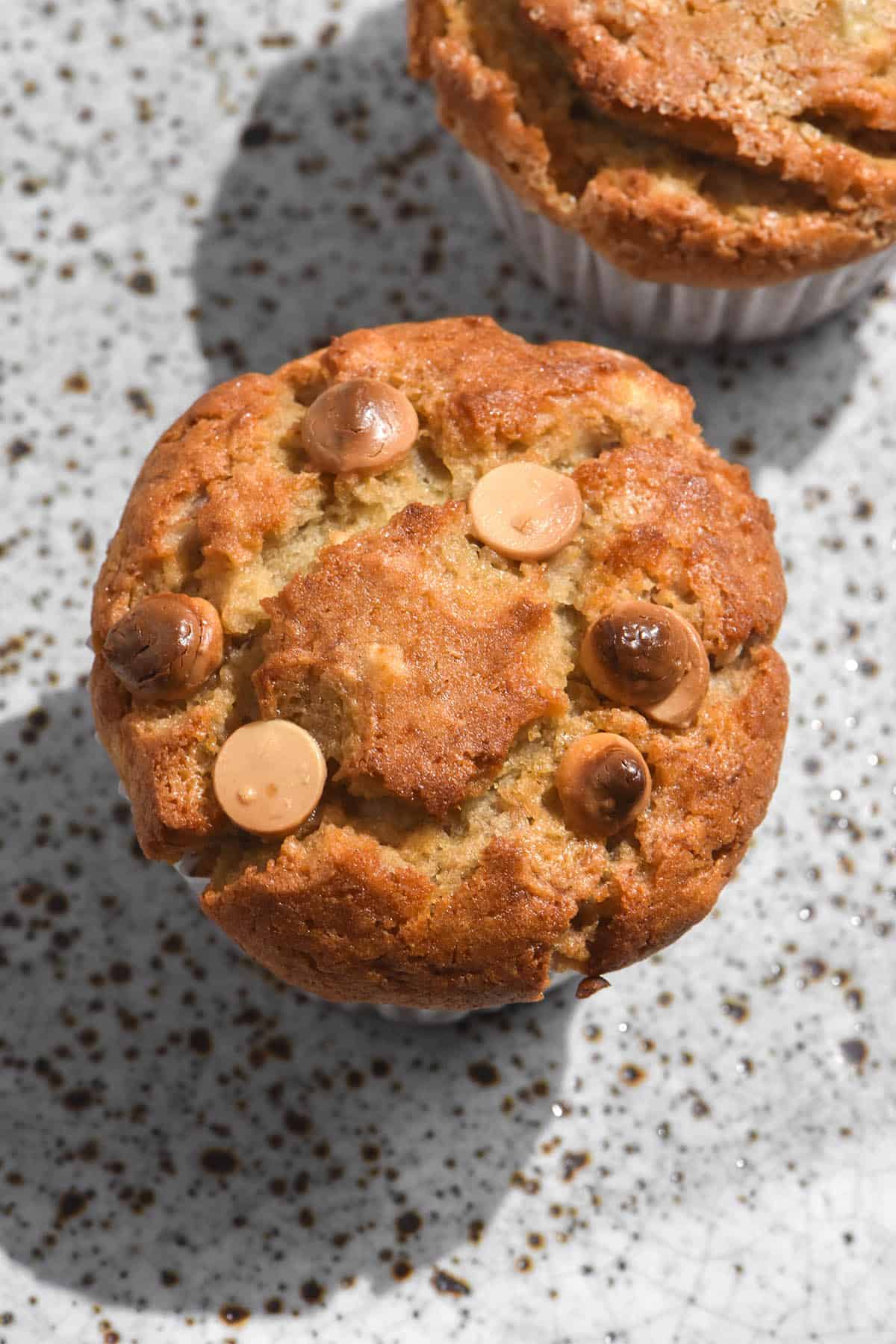 A close up macro image of two gluten free banana choc chip muffins on a white ceramic speckled plate. The muffins are golden brown and topped with either choc chips or finishing sugar. 