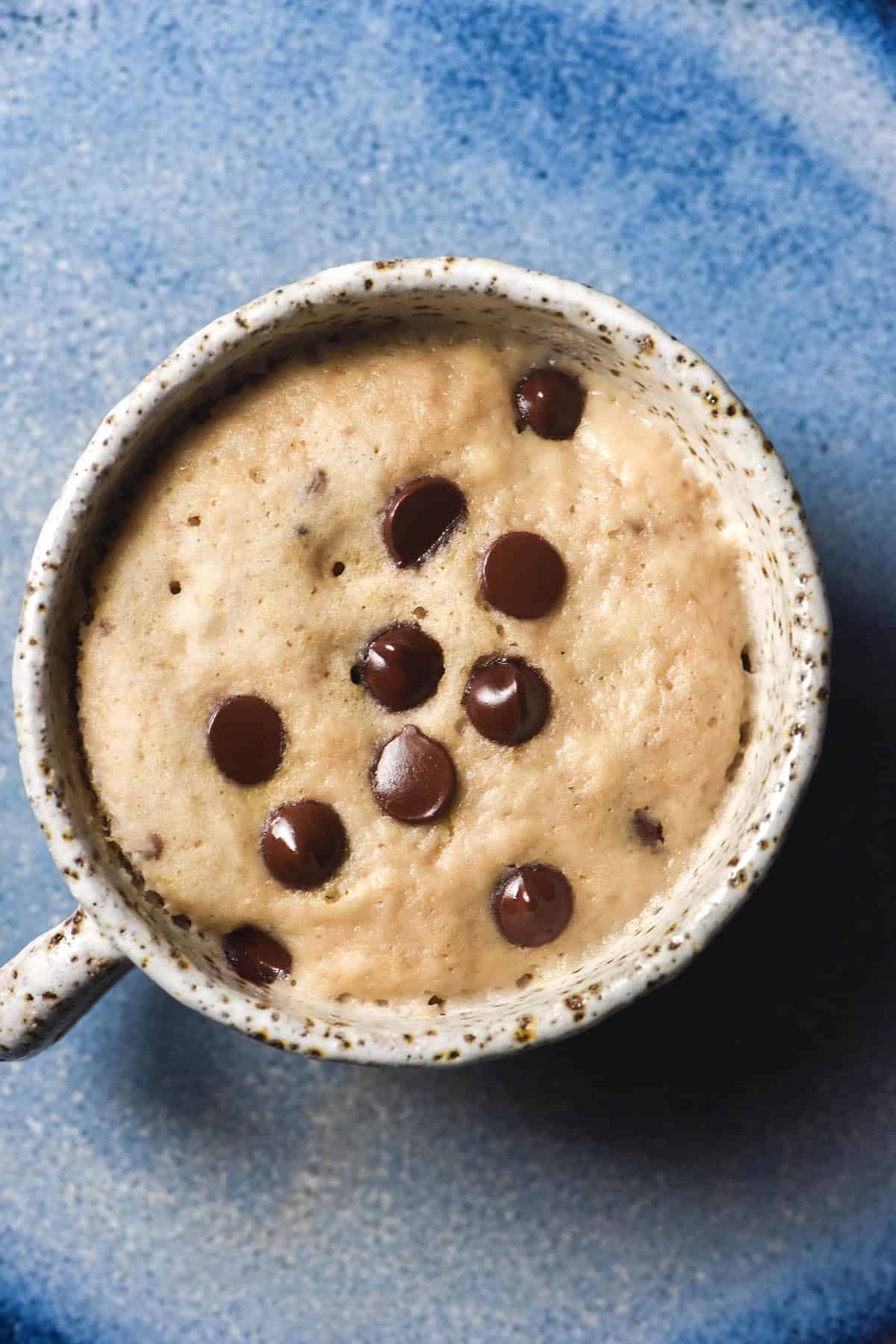 An aerial image of a gluten free banana mug cake in a white speckled ceramic mug on a bright blue ceramic plate