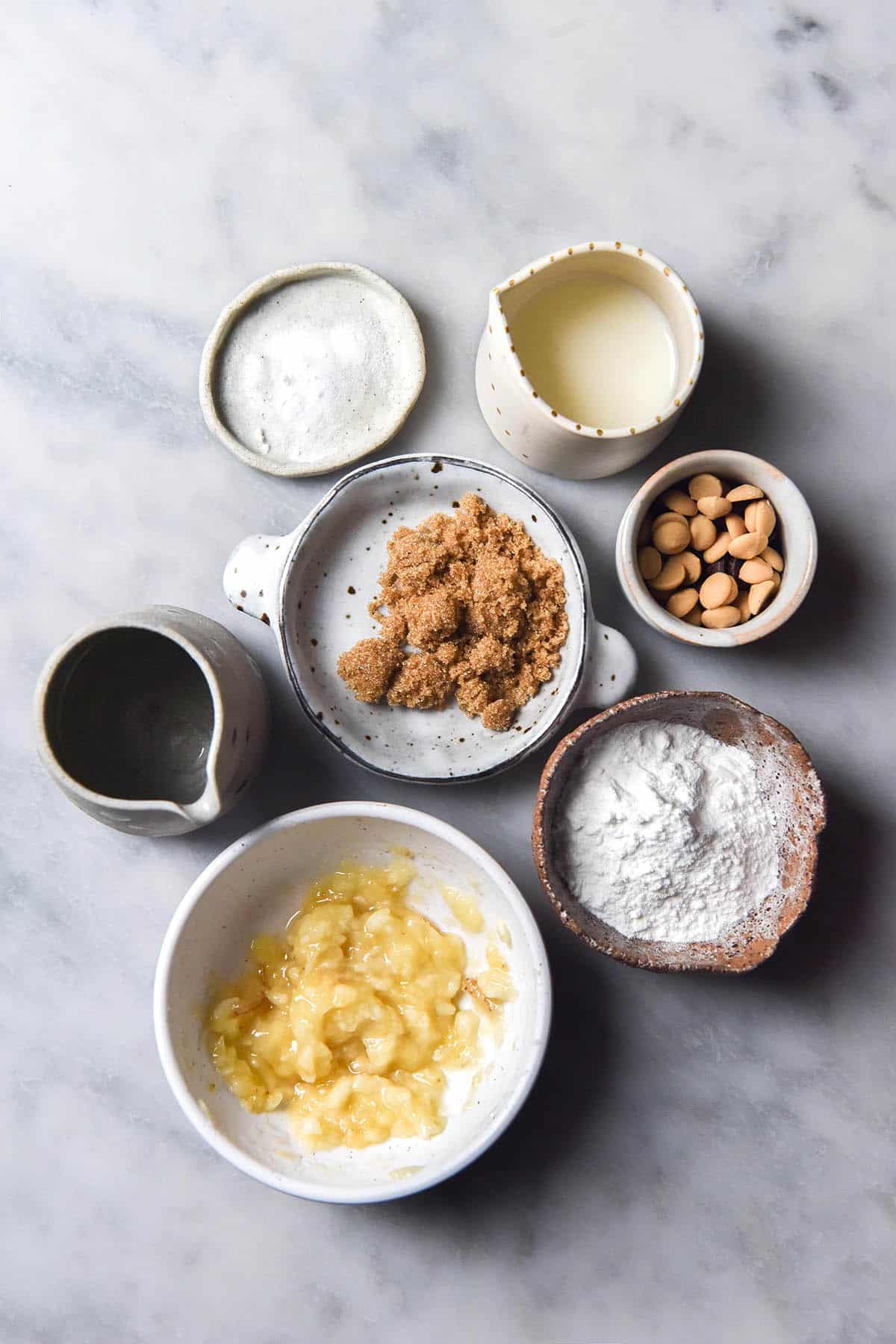 An aerial image of the ingredients used to make a gluten free banana mug cake arranged in white ceramic bowls on a white marble table