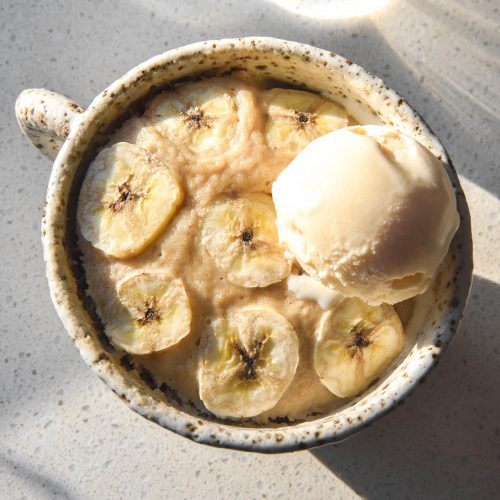 An aerial sunlit image of a gluten free banana mug cake in a white ceramic mug on a white benchtop with a glass of water in the background. The mug cake is topped with thin slices of banana and a scoop of vanilla ice cream.