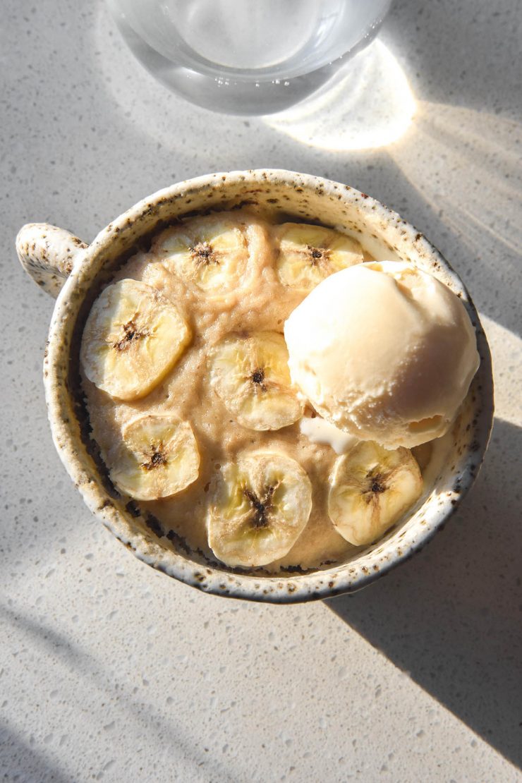 An aerial sunlit image of a gluten free banana mug cake in a white ceramic mug on a white benchtop with a glass of water in the background. The mug cake is topped with thin slices of banana and a scoop of vanilla ice cream.