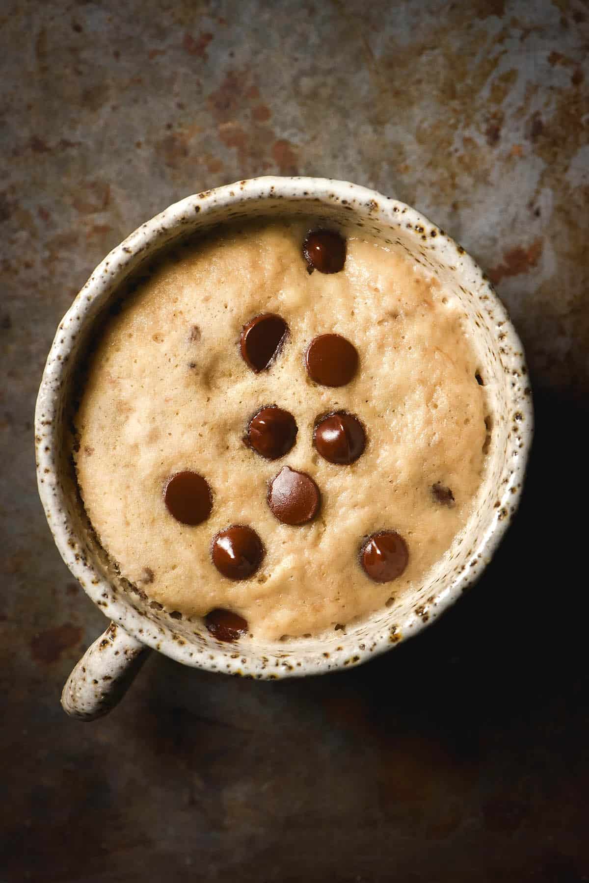 An aerial image of a gluten free banana choc chip mug cake in a white speckled ceramic mug on a dark steel backdrop.