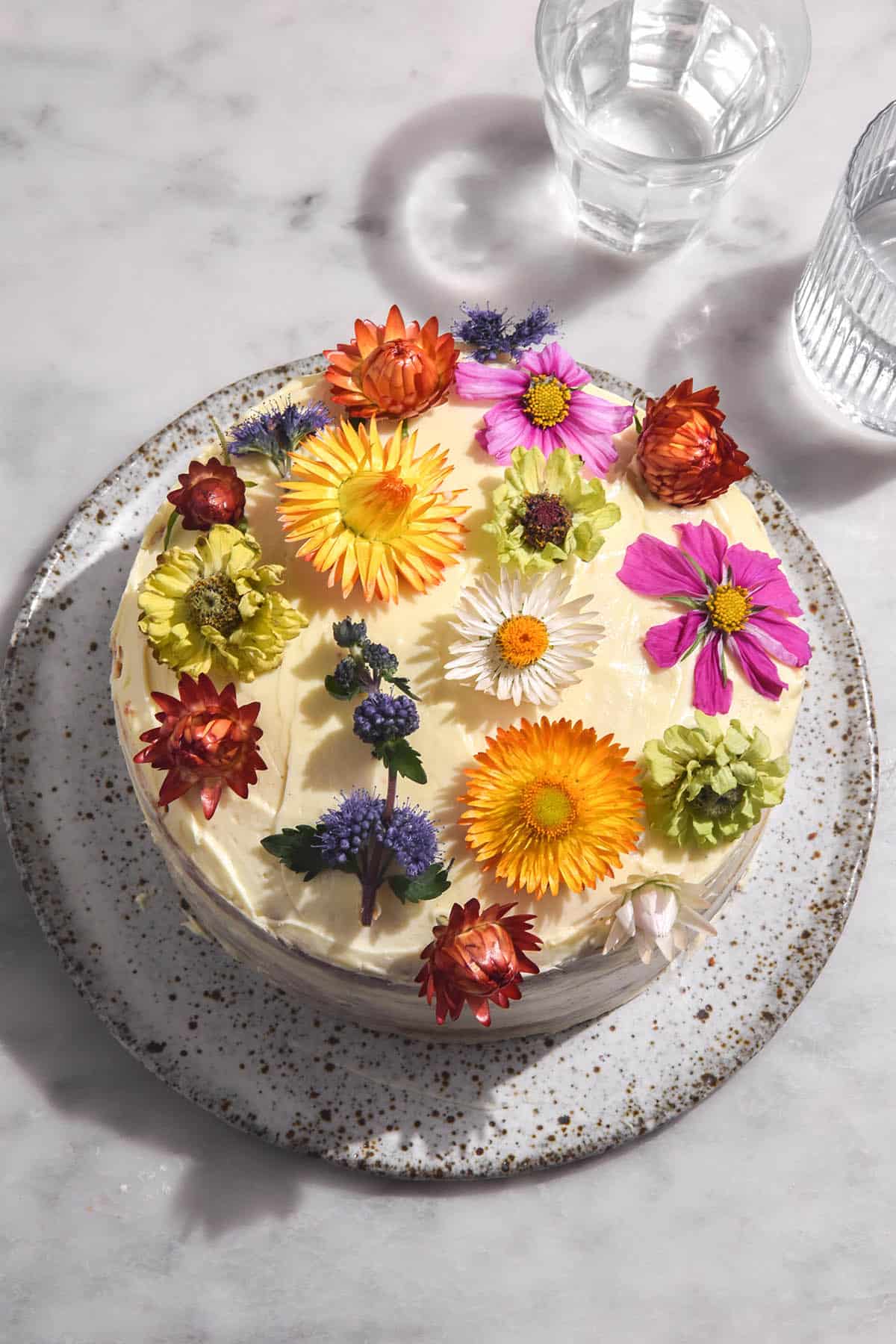 An aerial image of a gluten free cake decorated with lactose free cream cheese icing and multi coloured flowers. The cake sits on a white marble table and two glasses of water sit to the right of the cake. 