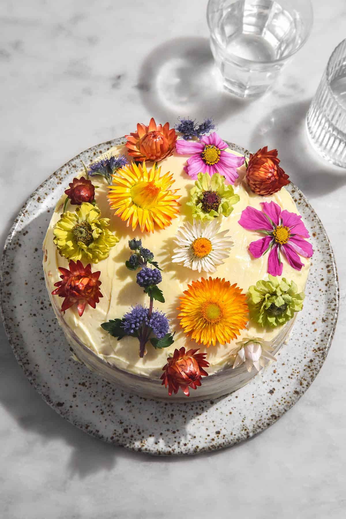 An aerial image of a gluten free cke filled with passion fruit curd and topped with cream cheese icing. The icing has been topped with brightly coloured fresh flowers and the cake sits on a white speckled ceramic plate atop a white marble table.