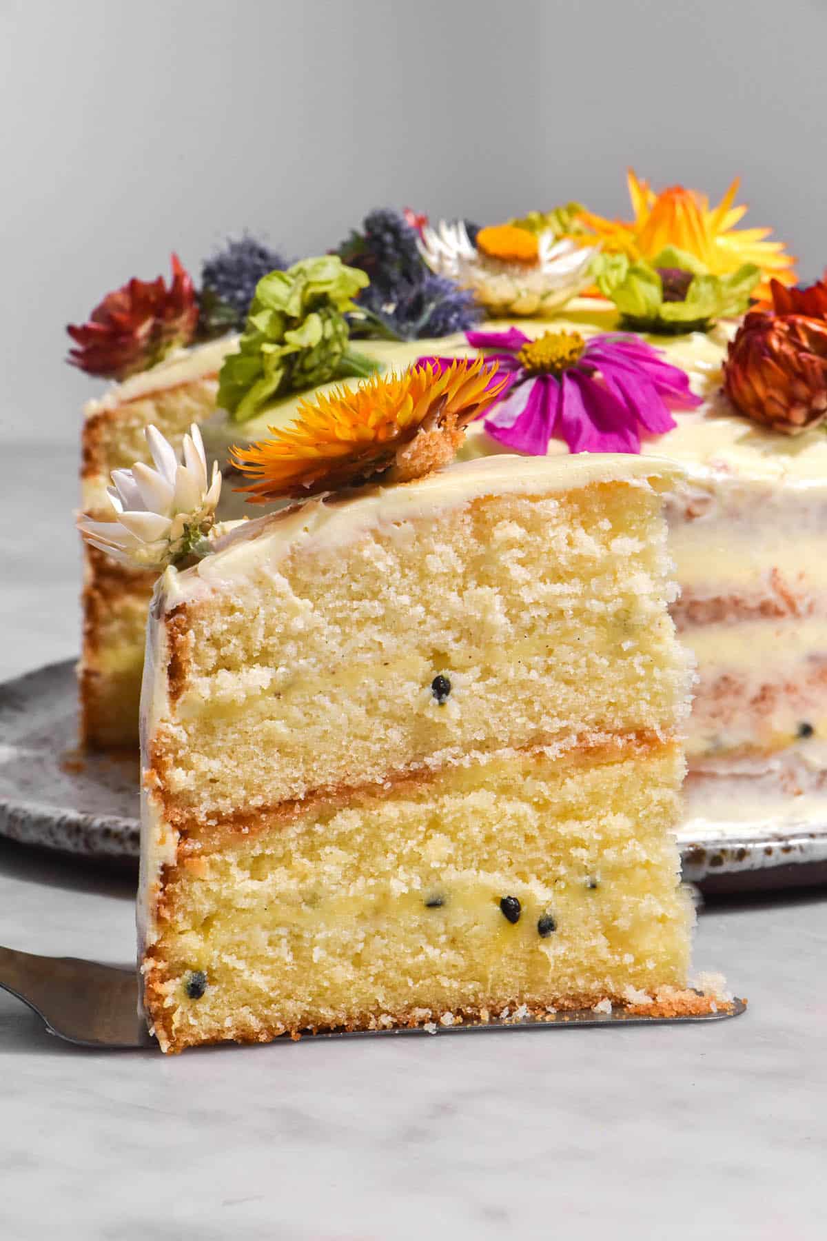 A side on view of a slice of gluten free cake with dairy free passion fruit curd layers and cream cheese icing. The cake is topped with multi coloured flowers and sits on a white marble table. 